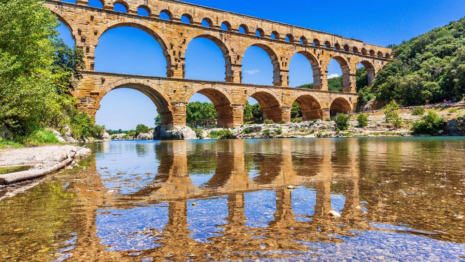 A stone aqueduct over a clear river with reflections, surrounded by greenery.