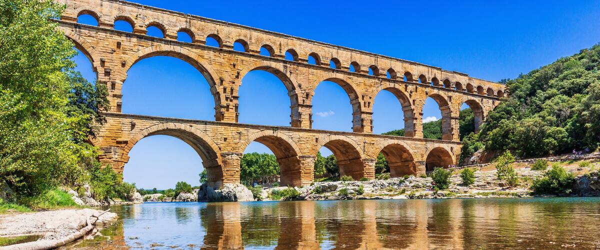A stone aqueduct over a clear river with reflections, surrounded by greenery.