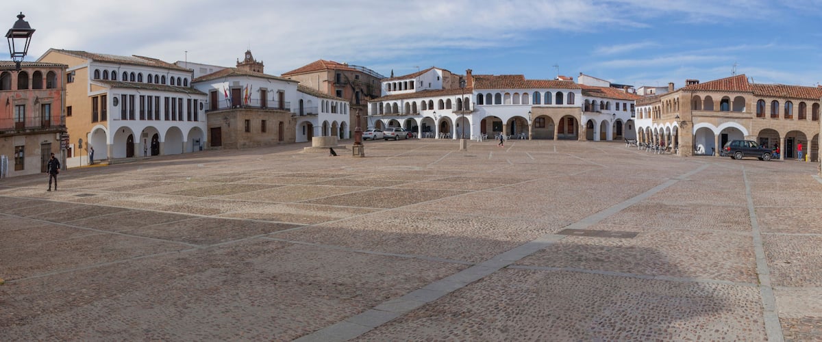 A historic plaza with cobblestone pavement, white buildings with arches, and a clear blue sky.