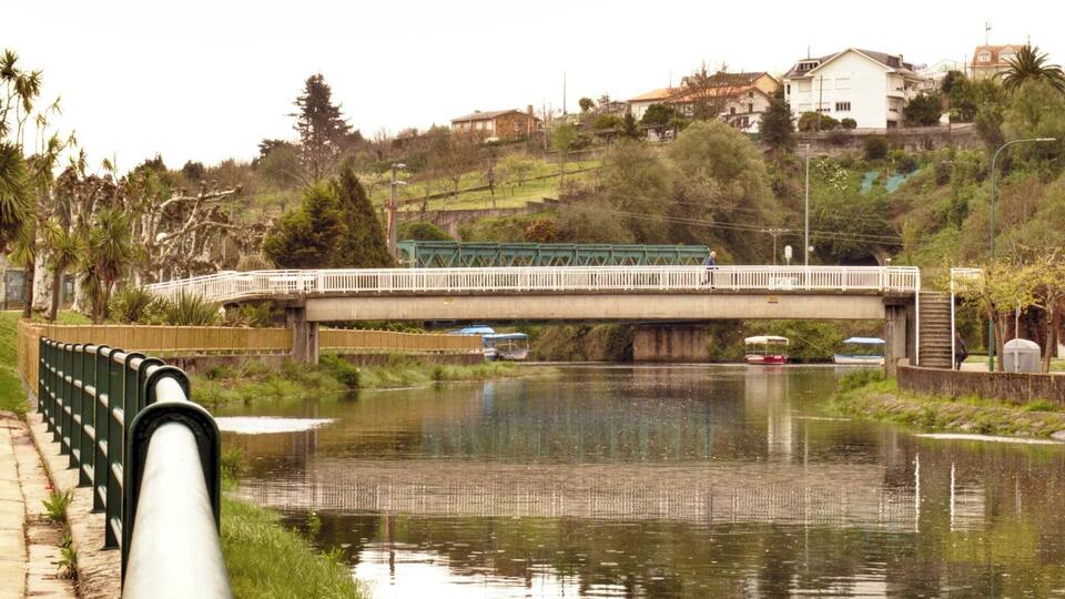 Un río con un puente, barcos y casas al fondo.