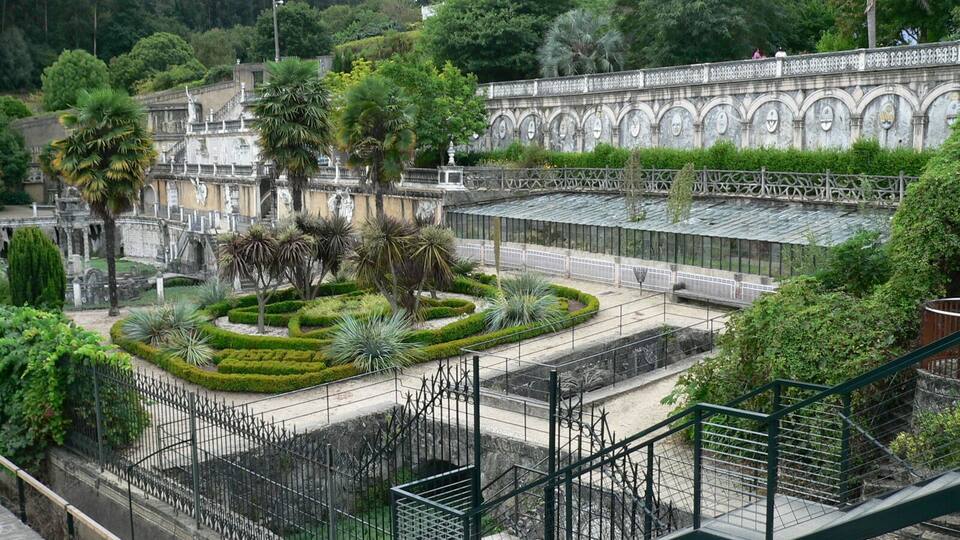 Un jardín ajardinado con una fuente, palmeras y un muro de piedra con arcos.