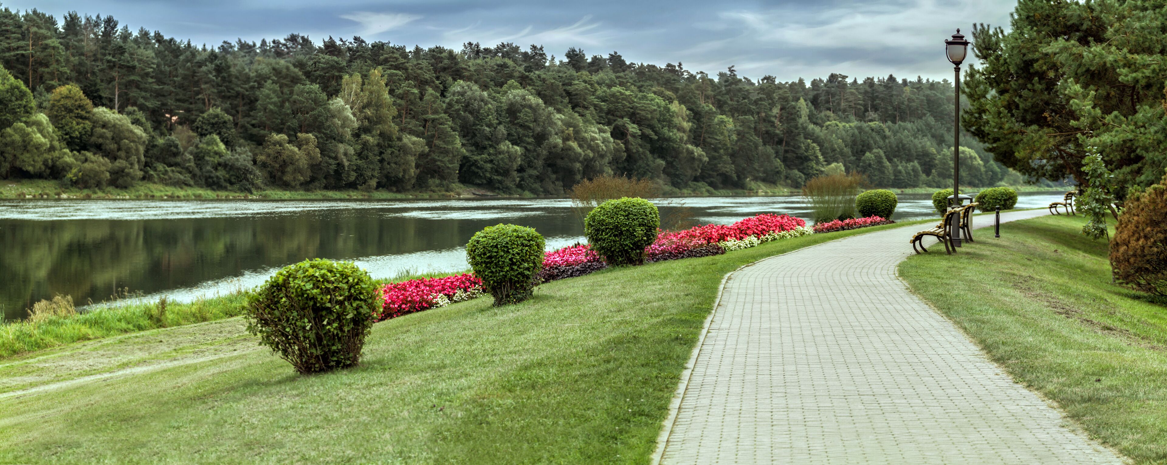A paved pathway alongside a river, bordered by lush greenery and flowering plants.