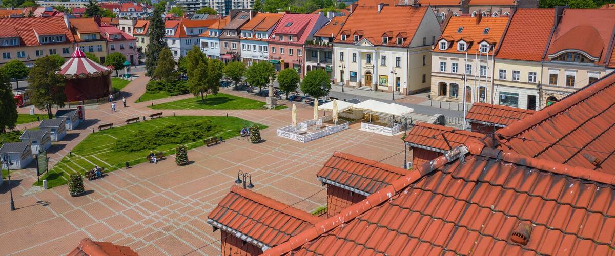 A town square with red-roofed buildings, a prominent church tower, and a central open space with benches and trees.
