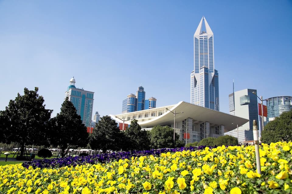 A cityscape with modern skyscrapers, a distinctive white building with a curved roof, and a field of yellow flowers in the foreground.