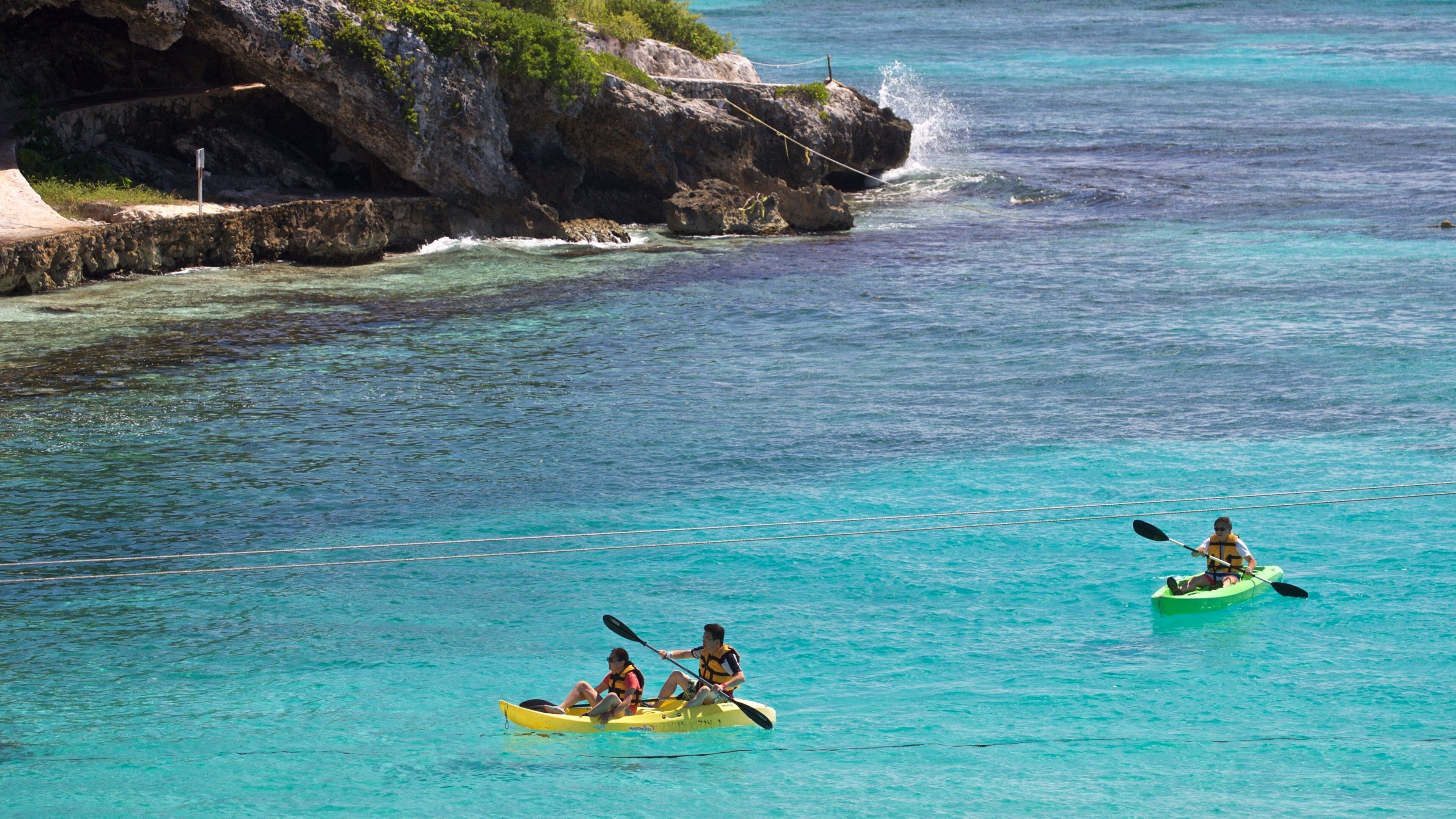 Two kayakers in a turquoise sea with rocky cliffs in the background.