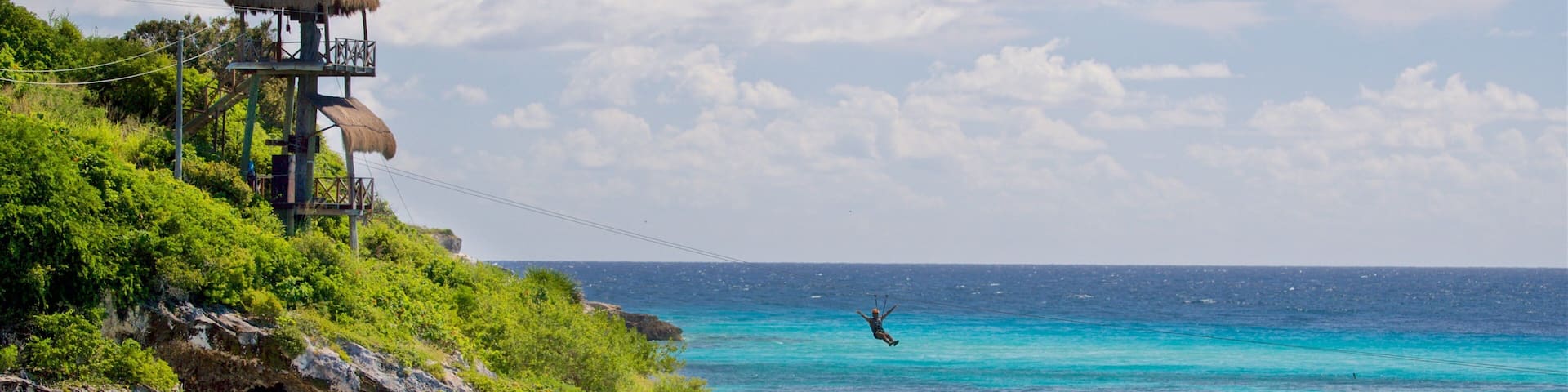 A coastal scene with a thatched-roof structure, clear turquoise waters, kayakers, and a person bungee jumping.