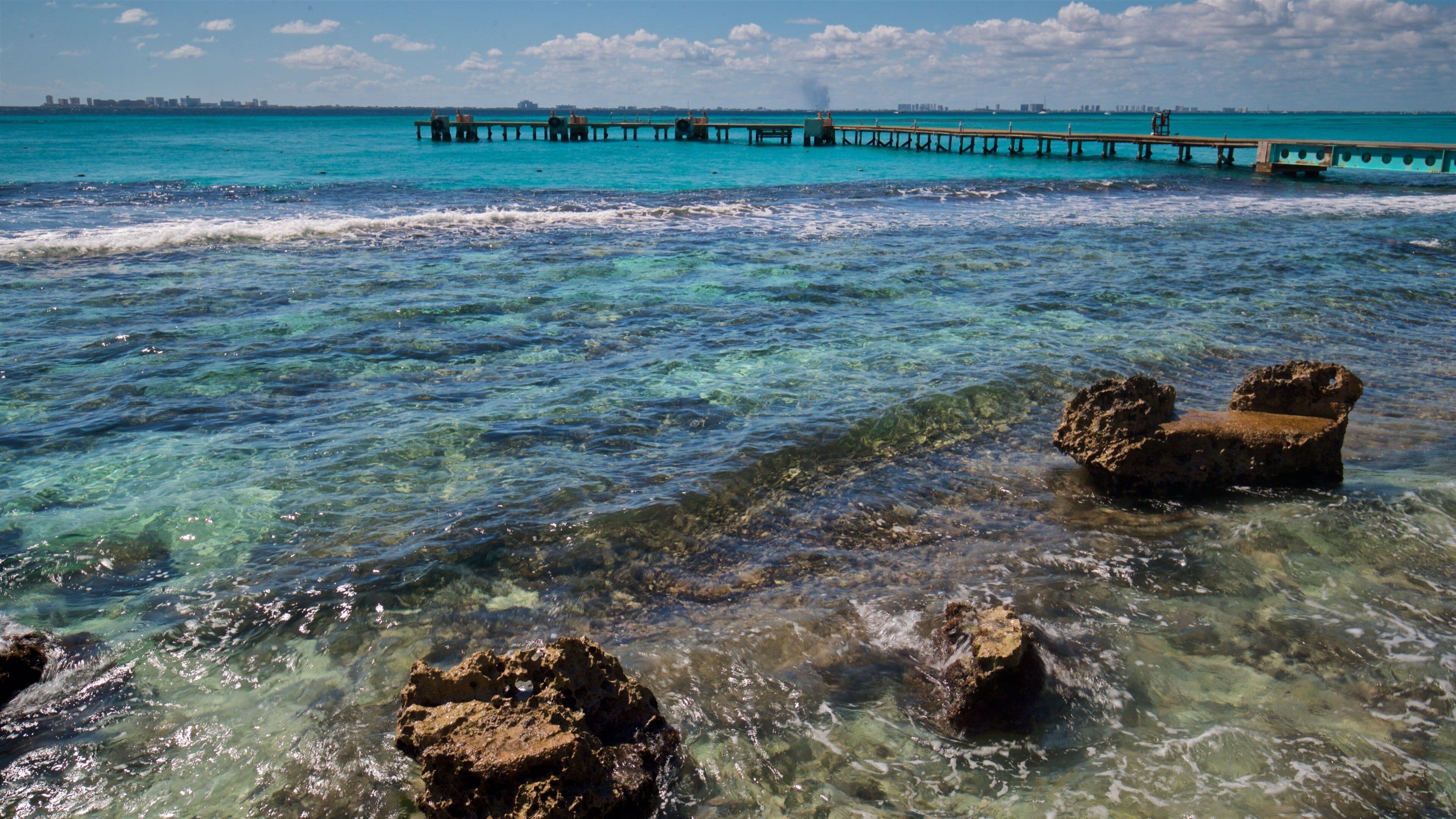 A clear turquoise sea with rocky formations in the foreground and a pier extending into the water in the background.
