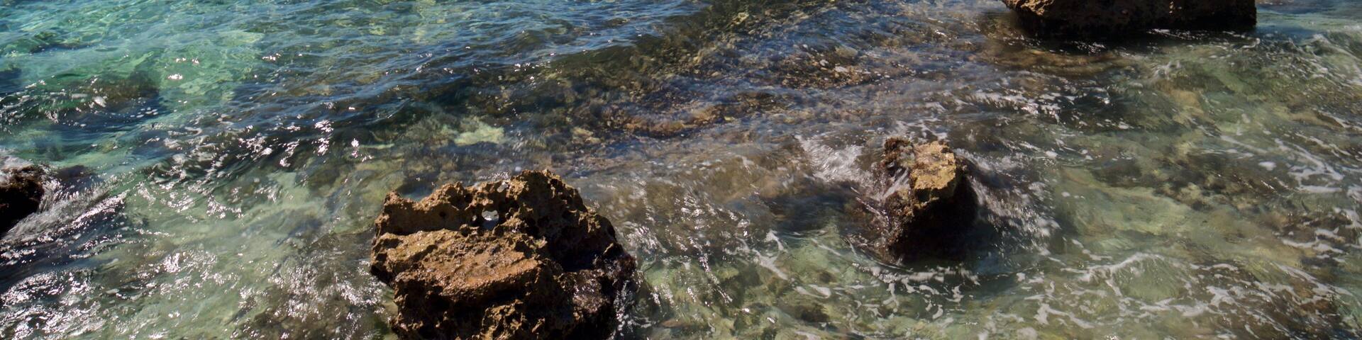 A clear turquoise sea with rocky formations in the foreground and a pier extending into the water in the background.