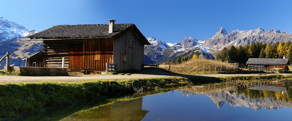 A wooden cabin by a calm lake with a mountain backdrop.