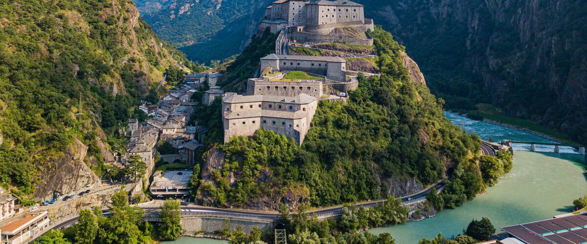 A historic fortress perched on a cliff overlooking a river, surrounded by lush greenery and steep mountains.