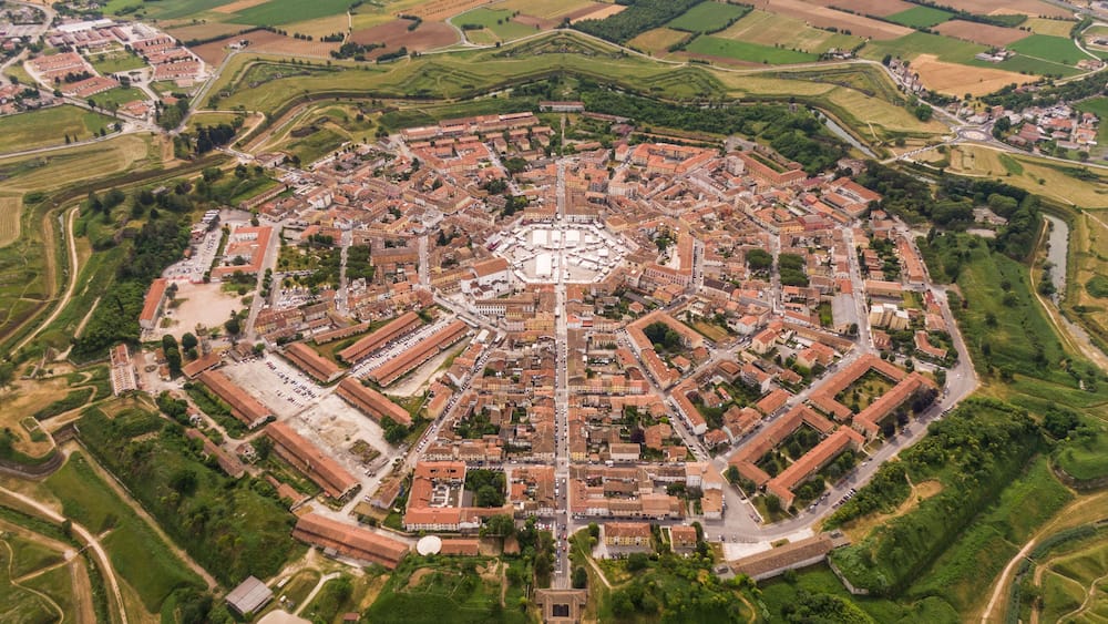 Ein Luftbild einer historischen, von Mauern umschlossenen Stadt mit einem zentralen Platz, umgeben von Ackerfeldern und einem Fluss.