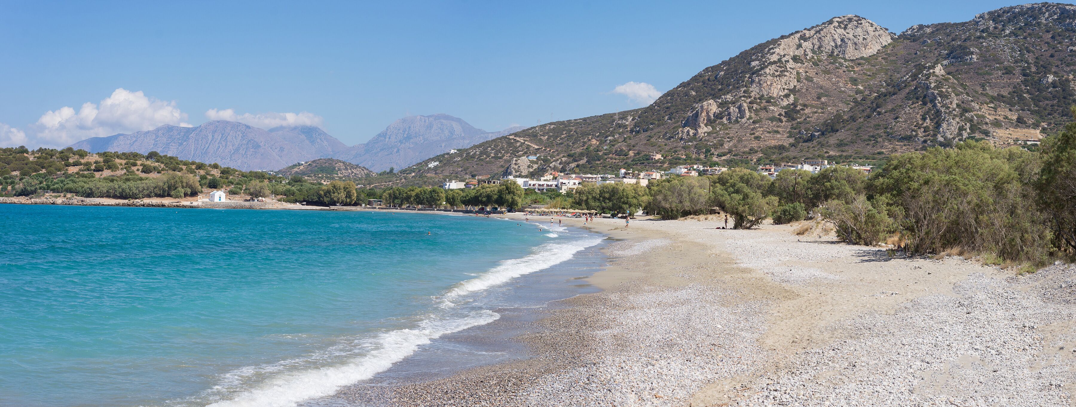 A pebble beach with clear turquoise waters, a white building, and mountains in the background.