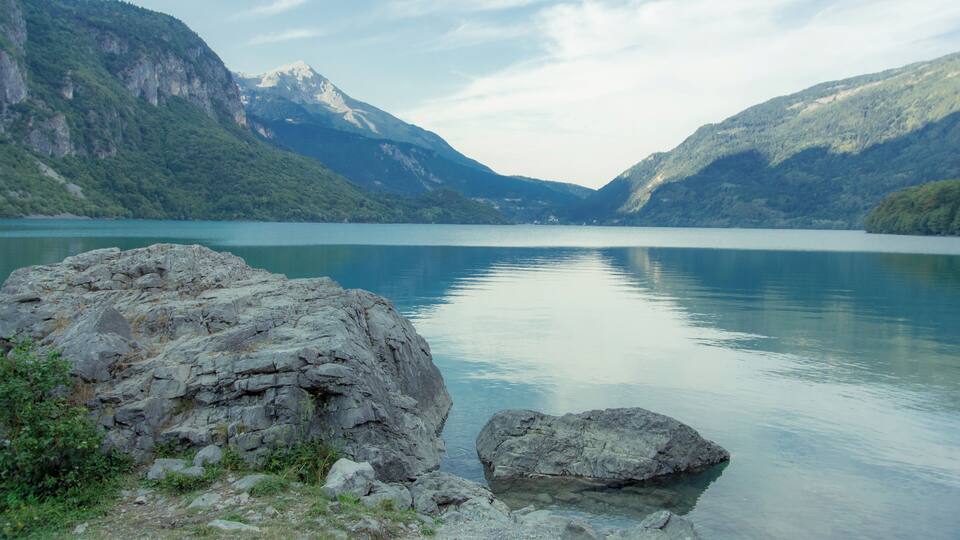 A calm lake surrounded by mountains with rocky outcrops in the foreground.
