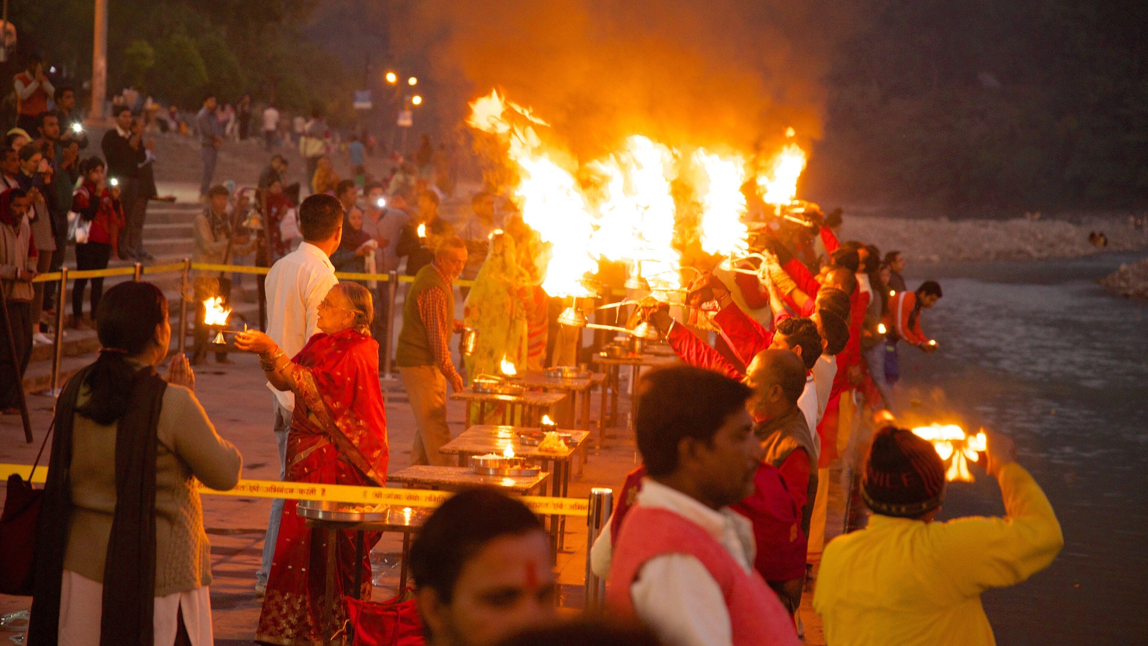 A nighttime outdoor gathering with people around a central fire, some holding candles, and a body of water in the background.