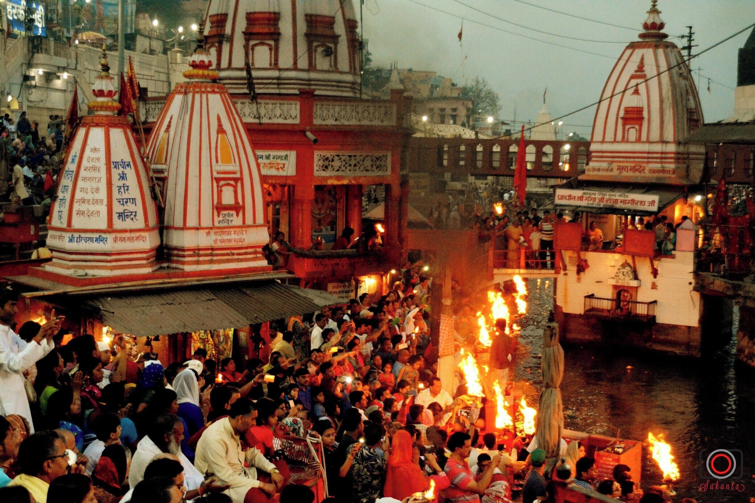 A crowded riverside with people and boats, surrounded by traditional buildings with domed structures.