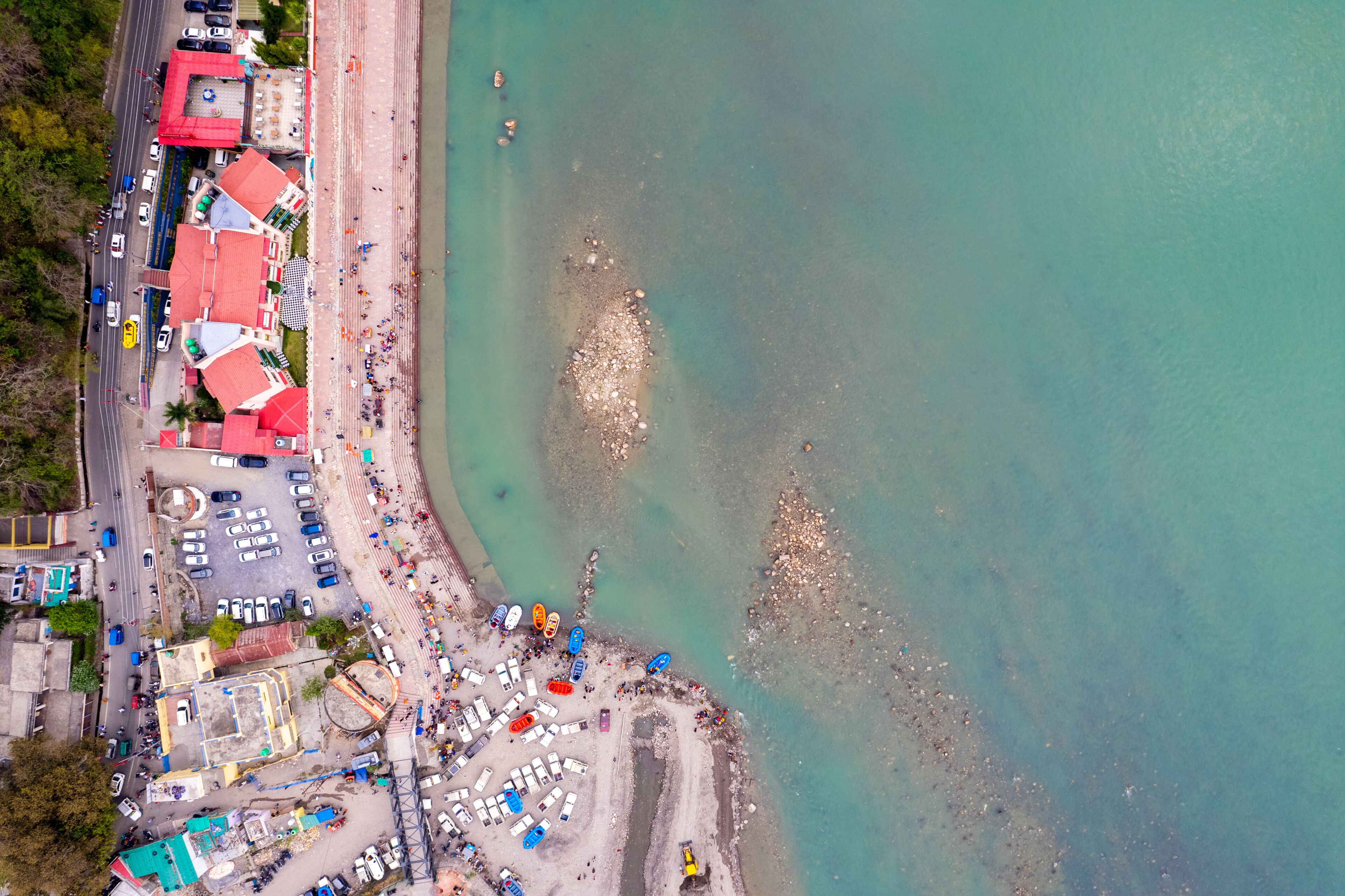 Vista aérea de uma área costeira com praia, veículos estacionados e edifícios com telhados vermelhos.