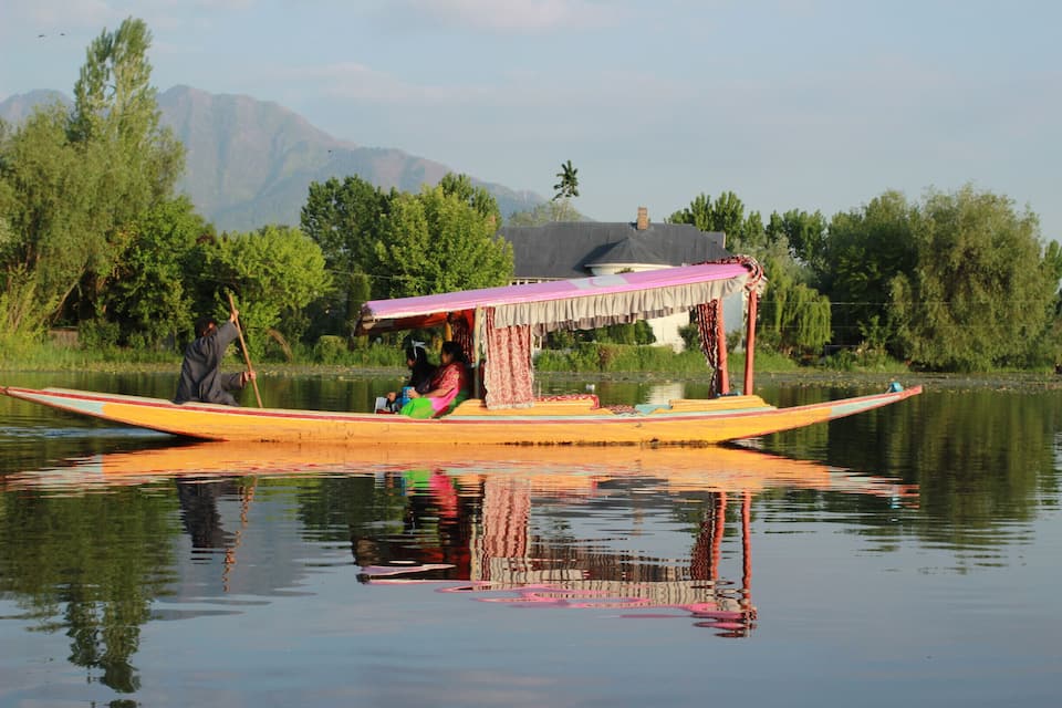 Um barco tradicional com toldo rosa em um rio calmo, cercado por vegetação exuberante e montanhas ao longe.