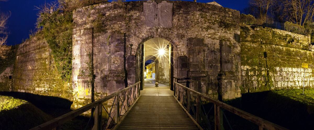 A stone archway leading to a building at night, surrounded by trees and a wooden walkway.