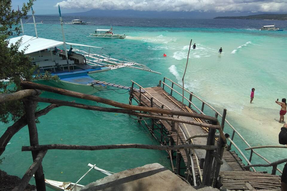 A coastal scene with a wooden pier leading to a sandy beach, boats in the water, and a mountainous landscape in the distance.