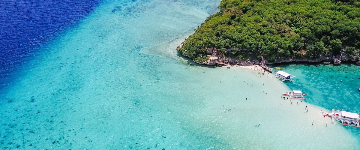 A turquoise beach with clear waters, a small island with dense greenery, and a few boats anchored near the shore.
