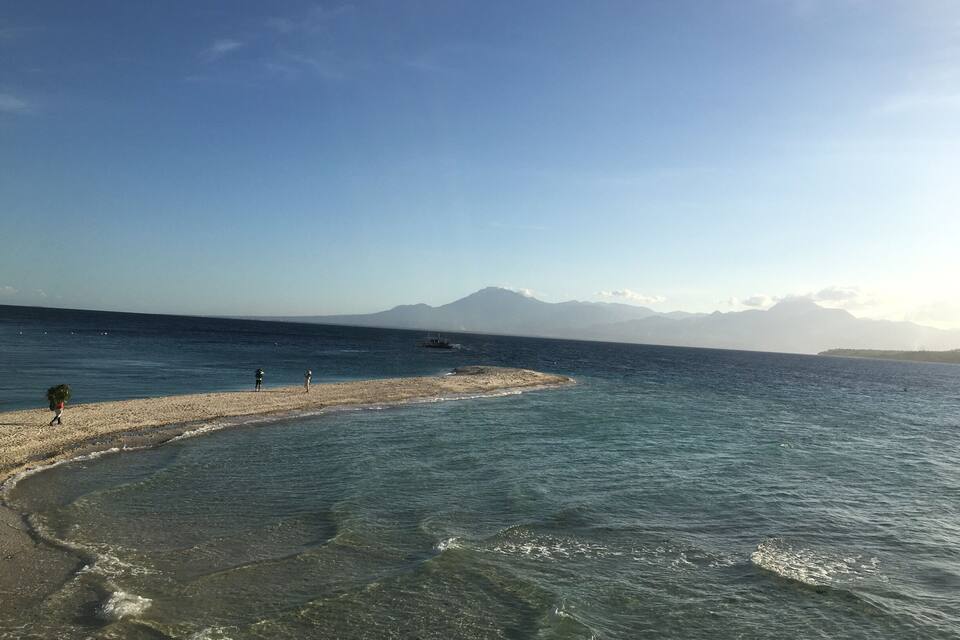 A beach with clear water, a small island, and mountains in the background.