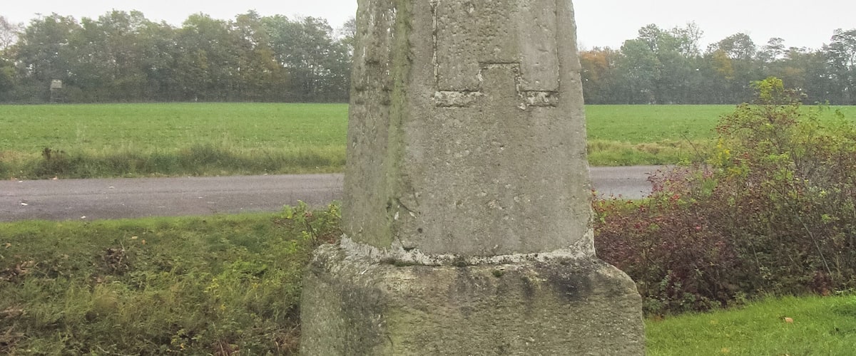 A tall, weathered stone monument with inscriptions, surrounded by grass and a rural landscape.