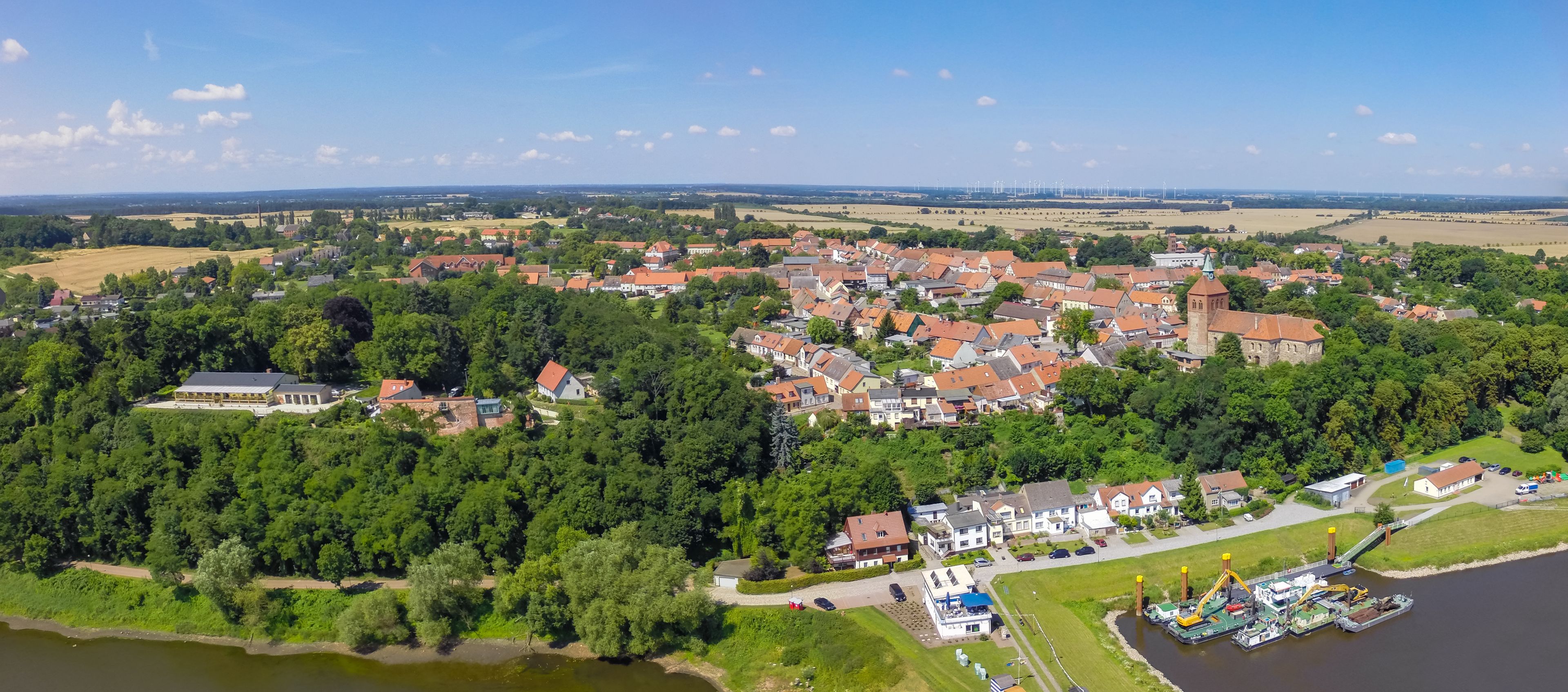 An aerial view of a town with a river, surrounded by greenery and wind turbines in the distance.
