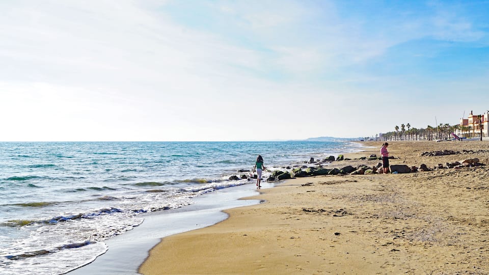 A sandy beach with waves, a clear sky, and buildings in the background.