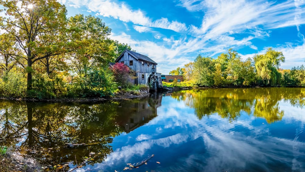 Un moulin en bois, situĂ© au bord dâune riviĂšre paisible, entourĂ© dâun feuillage automnal et surplombĂ© dâun ciel bleu clair.