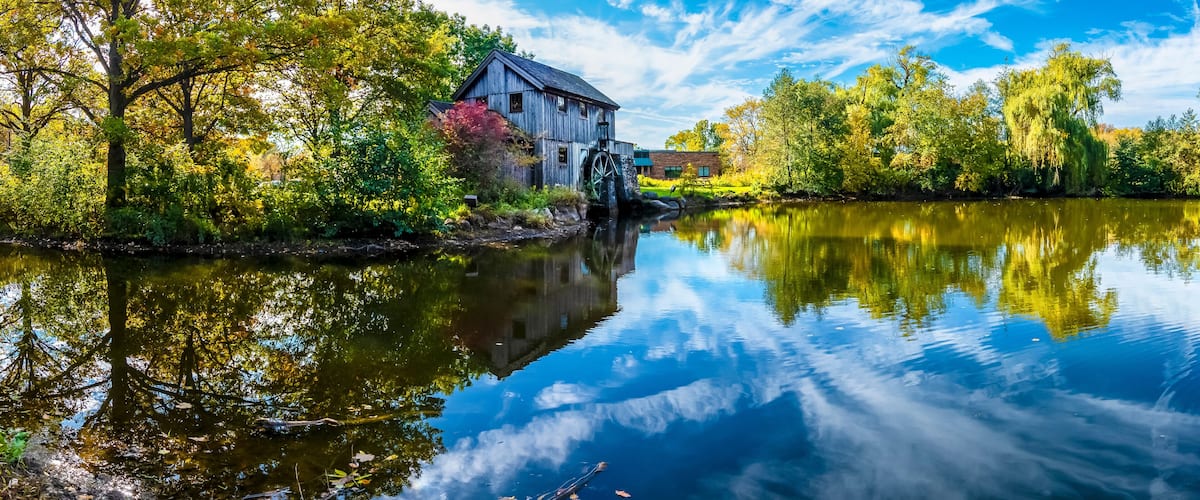 A wooden mill by a calm river, surrounded by autumn foliage and a clear blue sky.