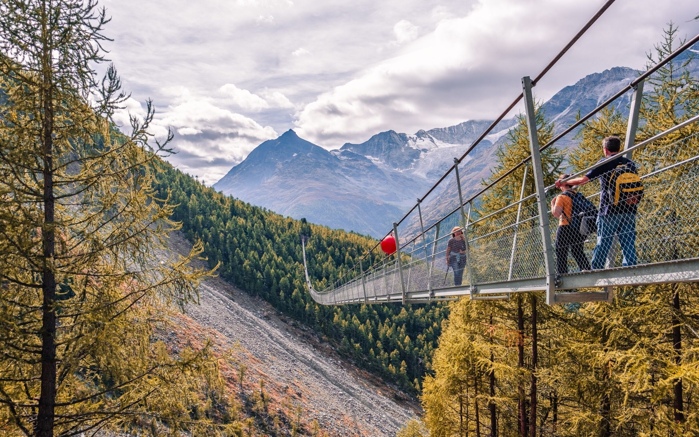 A suspension bridge with people walking across it, surrounded by autumn-colored trees and mountains in the background.