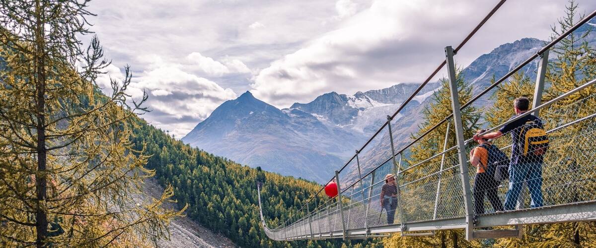 A suspension bridge with people walking across it, surrounded by autumn-colored trees and mountains in the background.
