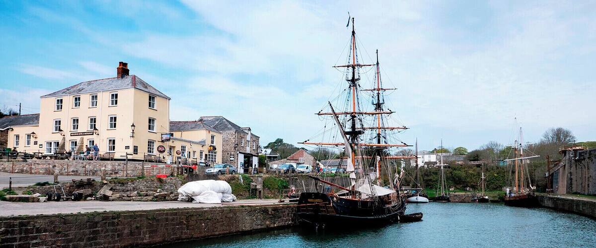 A tall ship docked at a harbor with a building and a stone wall in the background.