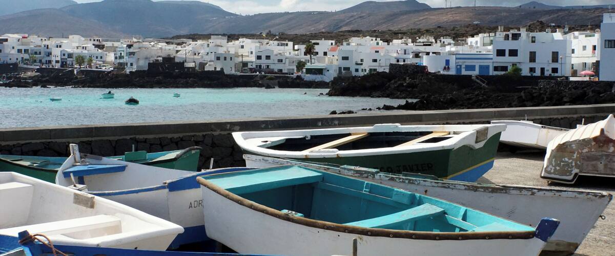 A coastal village with white buildings, boats docked at the pier, and mountains in the background.