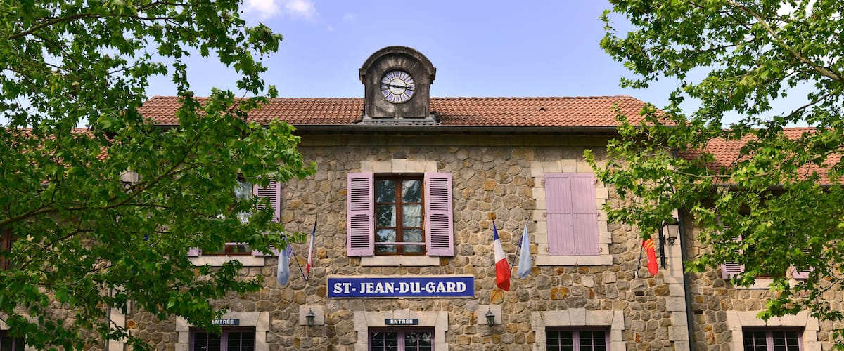A stone building with a clock, flags, and a sign reading "ST. JEAN-DU-GARD".