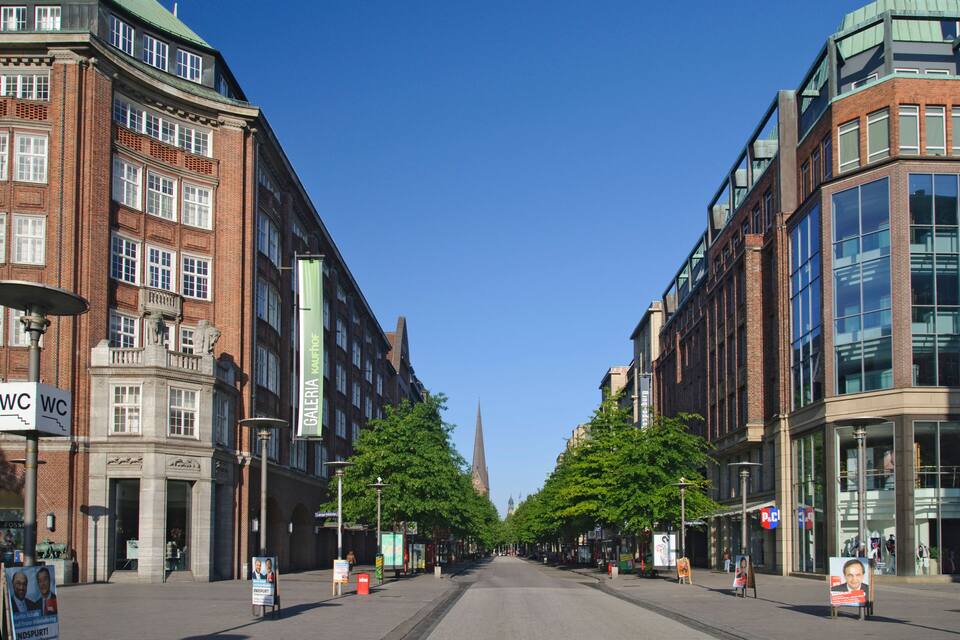 A city street lined with historic buildings, a prominent church spire in the distance, and a clear blue sky.