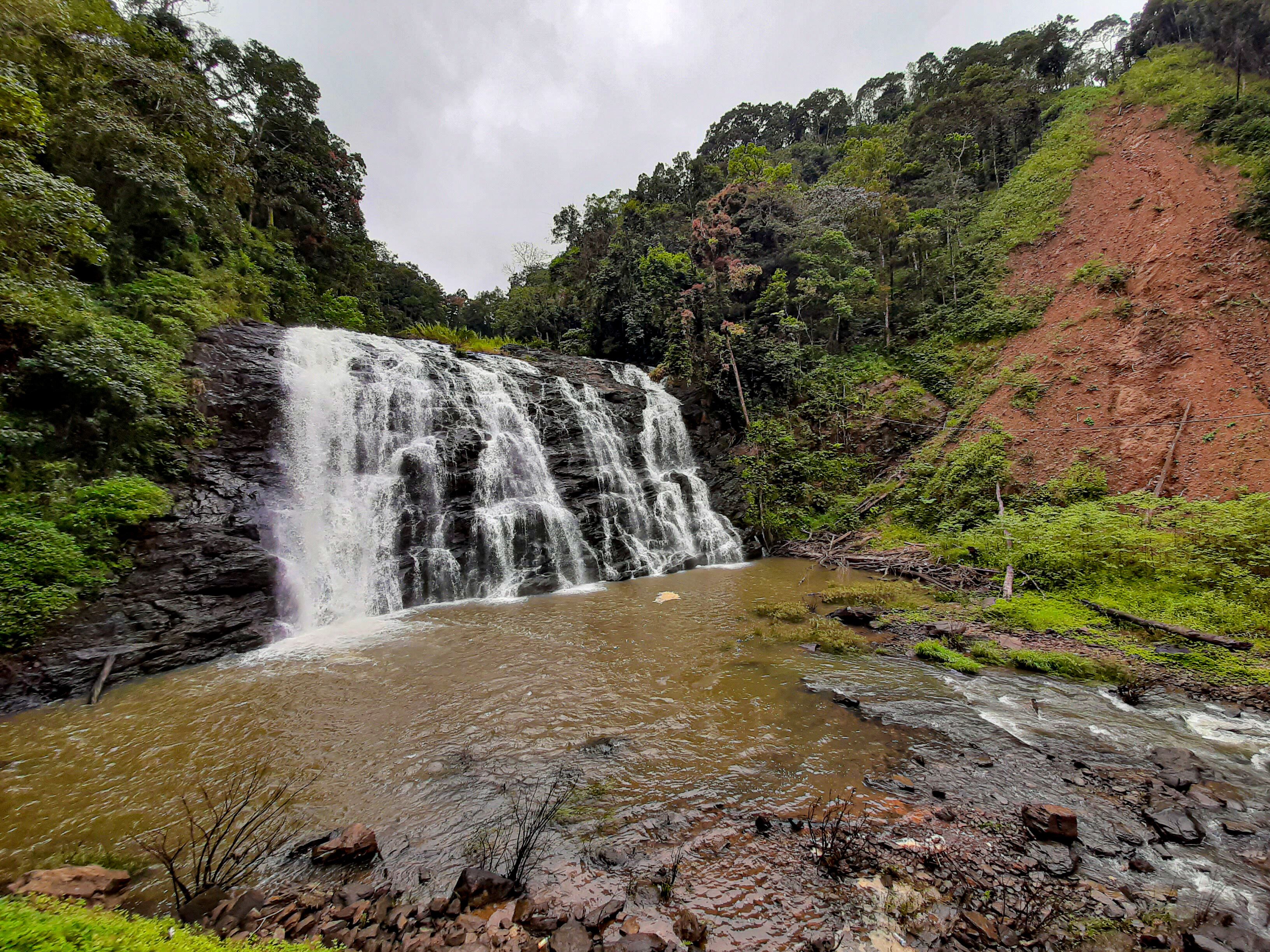 Une cascade se jetant dans une rivière, le tout entouré d’une végétation luxuriante et bordé d’une colline rougeoyante.