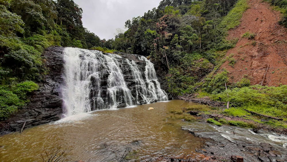 Ein Wasserfall stürzt in einen Fluss, umgeben von üppiger Vegetation und einem roten Hügelhang.