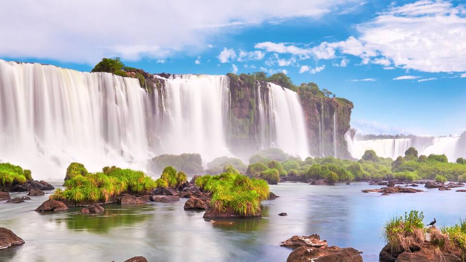 Iguazu waterfalls in Argentina. Panoramic view of many majestic powerful water cascades with mist and clouds. Panoramic image of Iguazu valley with grass and stones in calm water.