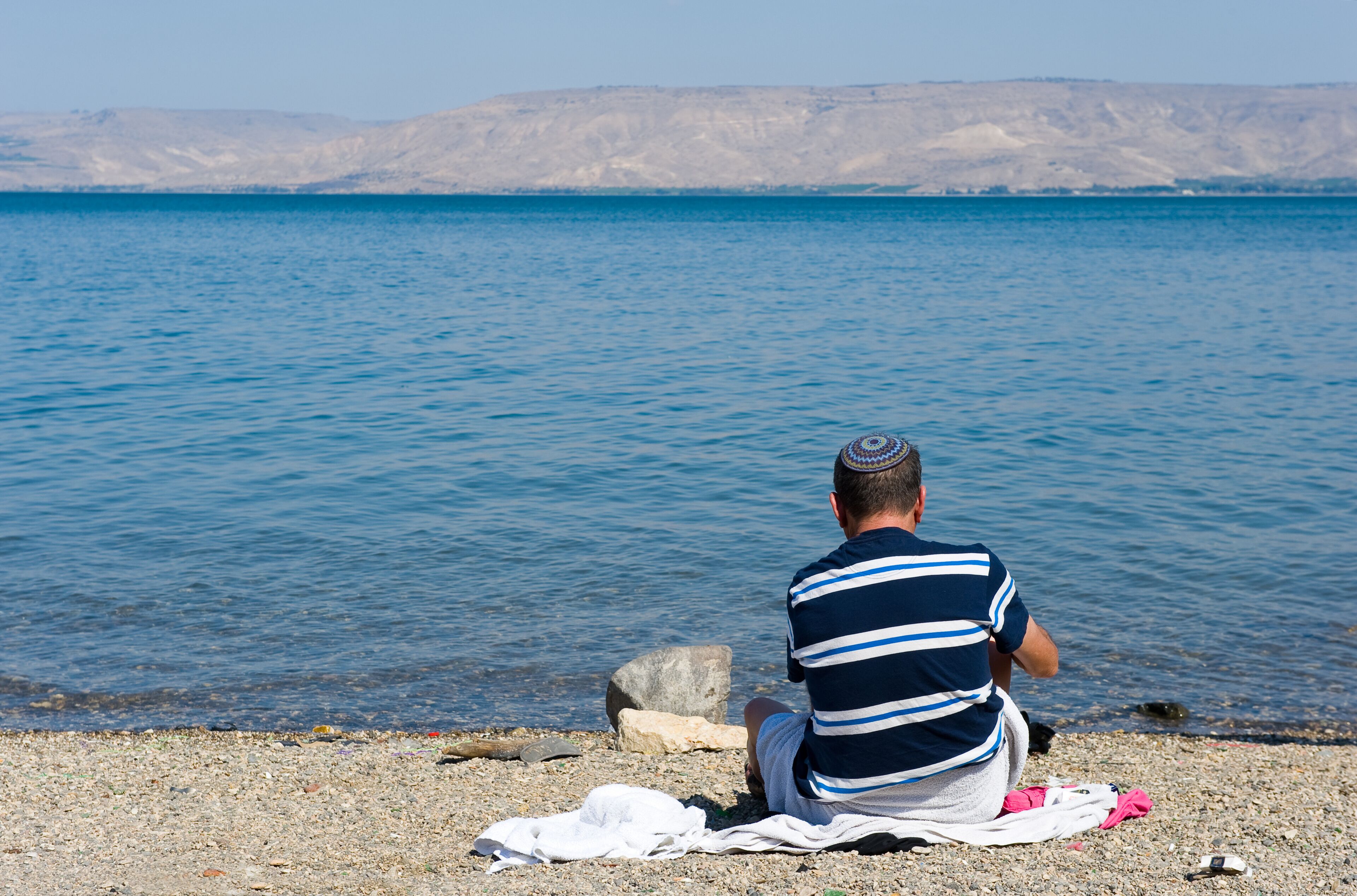 A man sitting on a pebble beach facing a calm sea with distant cliffs.
