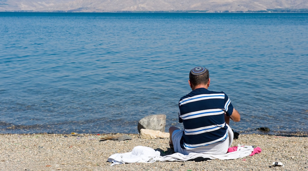 A man sitting on a pebble beach facing a calm sea with distant cliffs.
