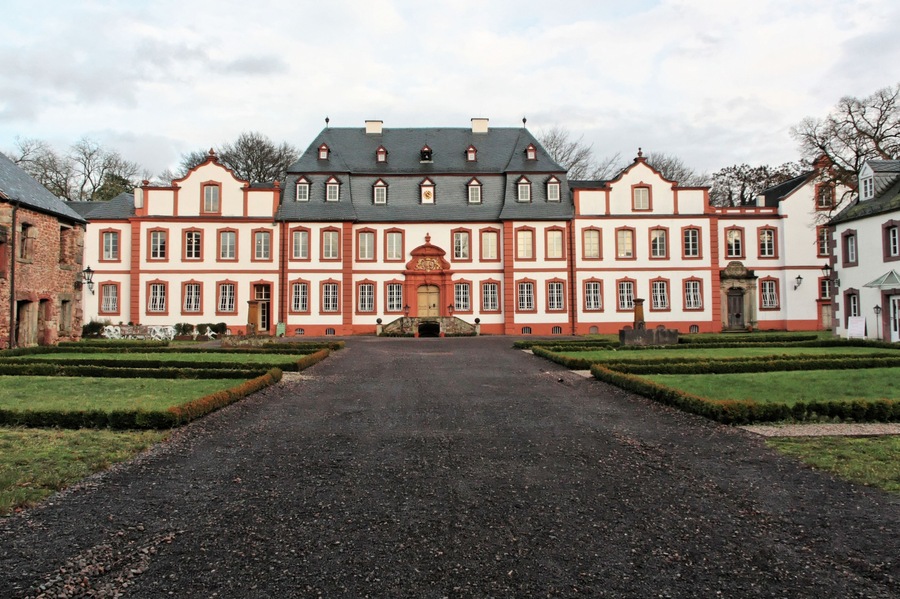 A large, historic building with a symmetrical facade, a central entrance, and a formal garden with trimmed hedges.
