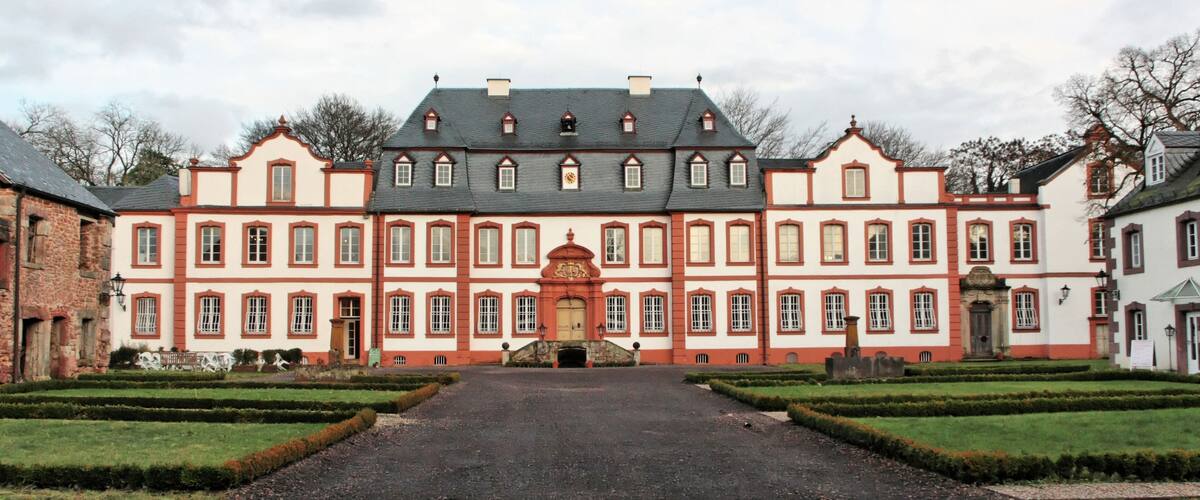 A large, historic building with a symmetrical facade, a central entrance, and a formal garden with trimmed hedges.