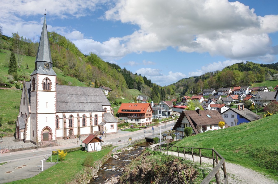 Une église dotée d’un clocher imposant, une petite chapelle et un quartier résidentiel avec des maisons et un ruisseau.