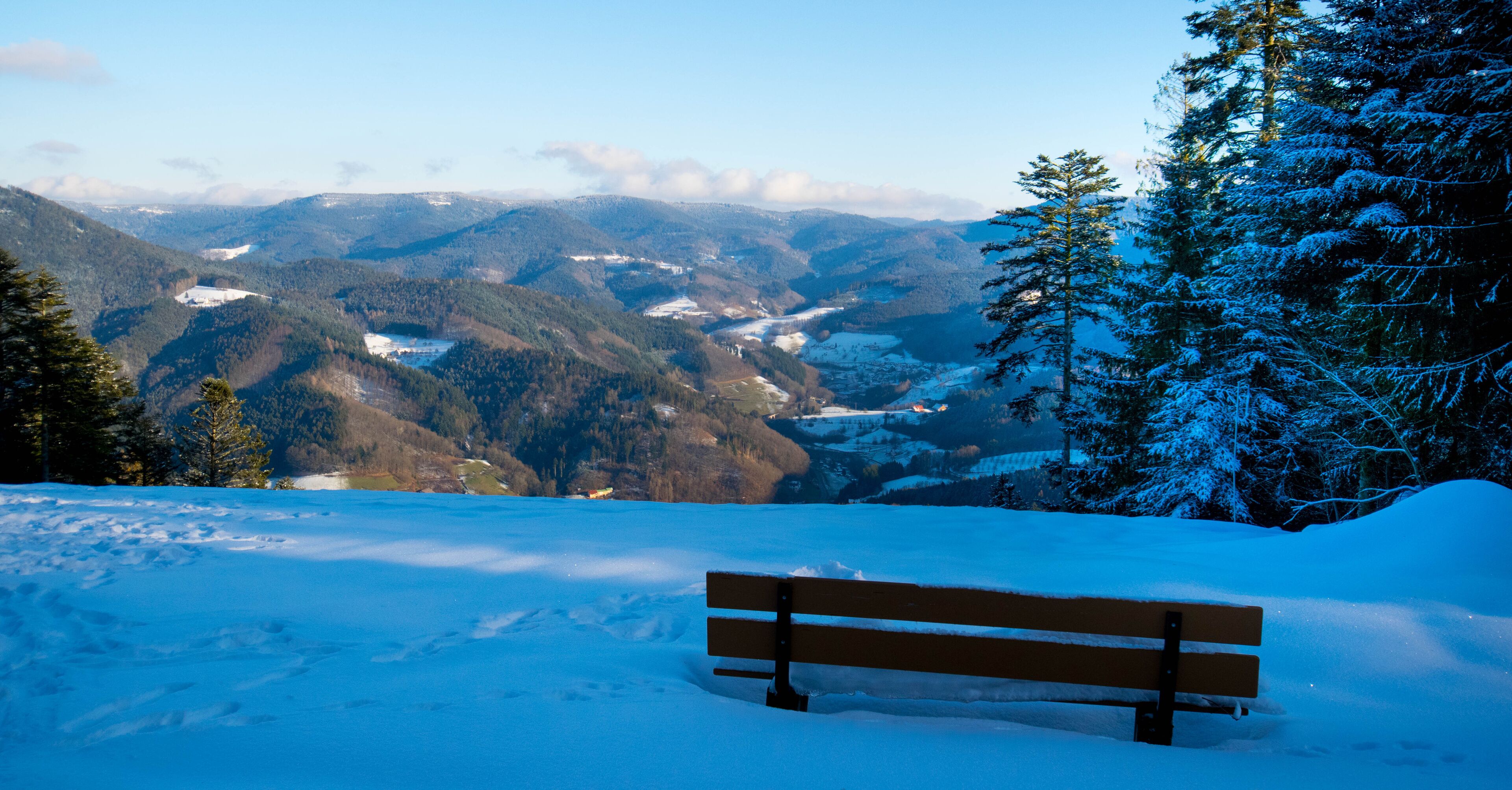 A snow-covered bench overlooking a mountainous landscape with a valley and snow-covered trees.