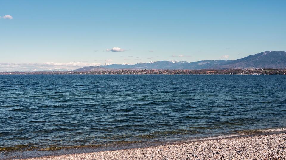 A lake with a pebble beach and a distant cityscape with mountains.