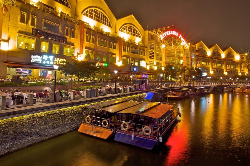 A riverside promenade at night with illuminated buildings, outdoor dining, and a boat docked by the water.