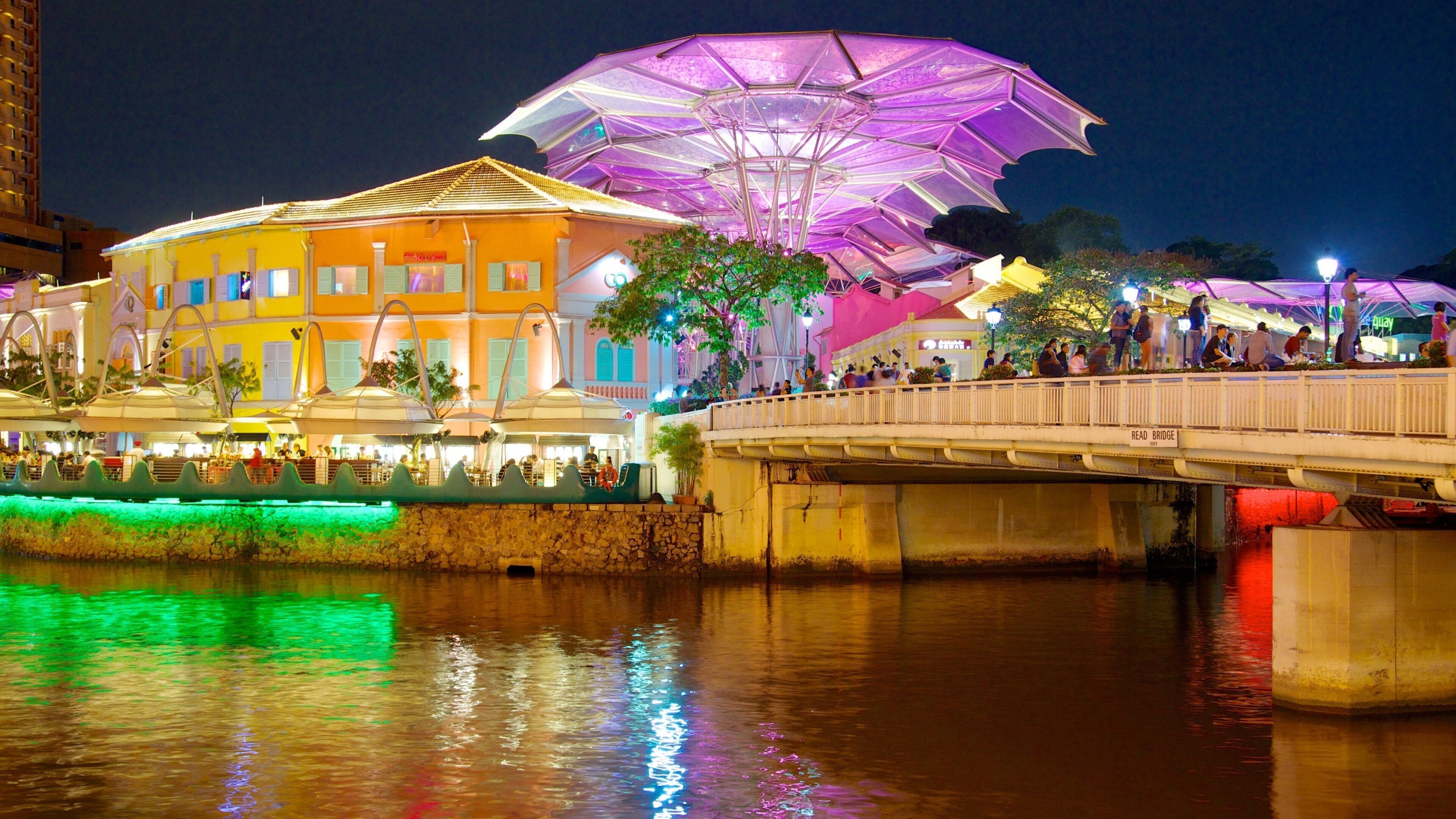 Un pont illuminé de mille feux, où l’on voit des gens qui le traversent, un bâtiment aux couleurs vives surmonté d’une grande structure en forme de parapluie, et des reflets sur l’eau en contrebas.