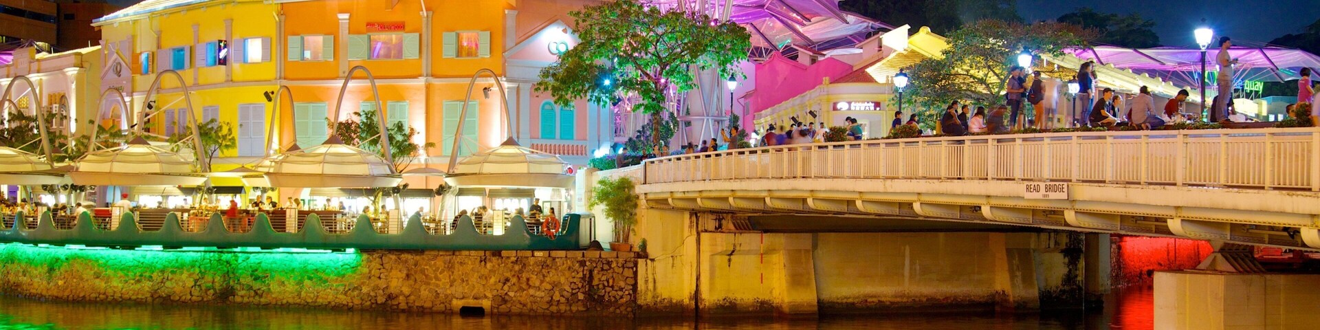 A brightly lit bridge with people walking across it, a colorful building with a large umbrella structure, and reflections on the water below.