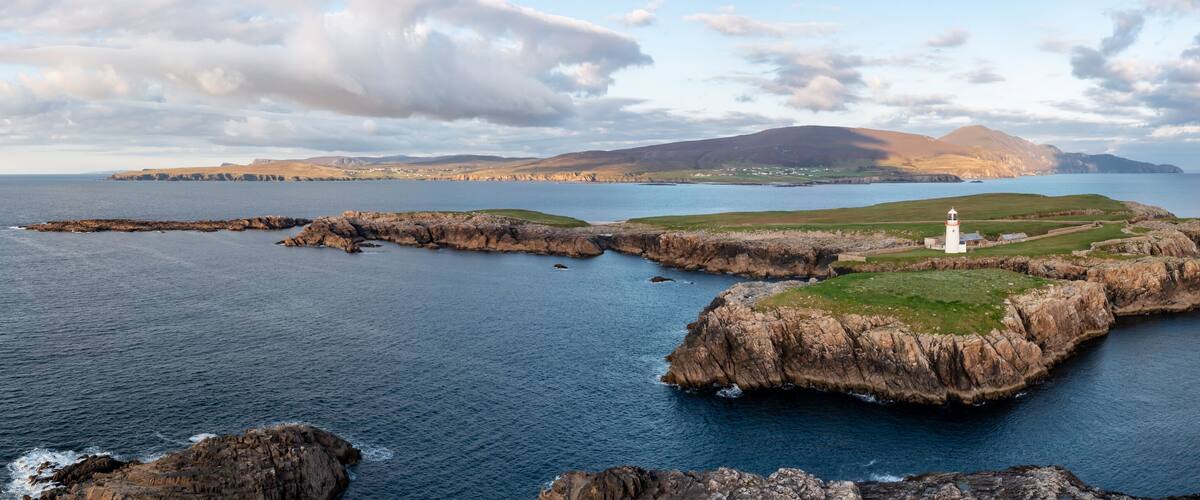 A rugged coastline with a lighthouse, rocky outcrops, and a distant mountain range.